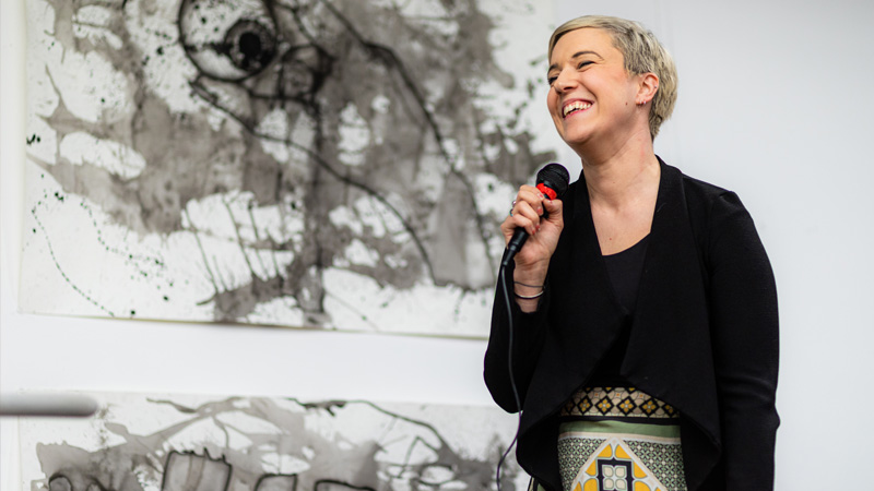 a smiling student speaks on microphone with artwork behind them