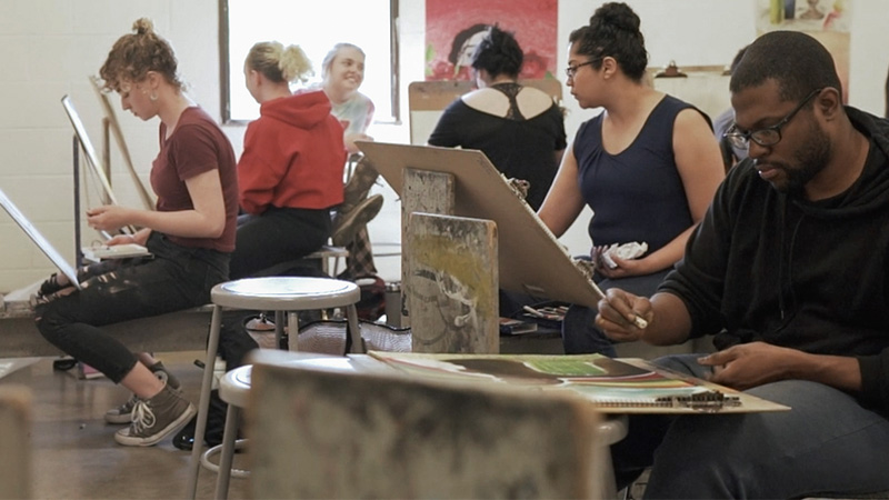 a group of students sit in class painting at easels