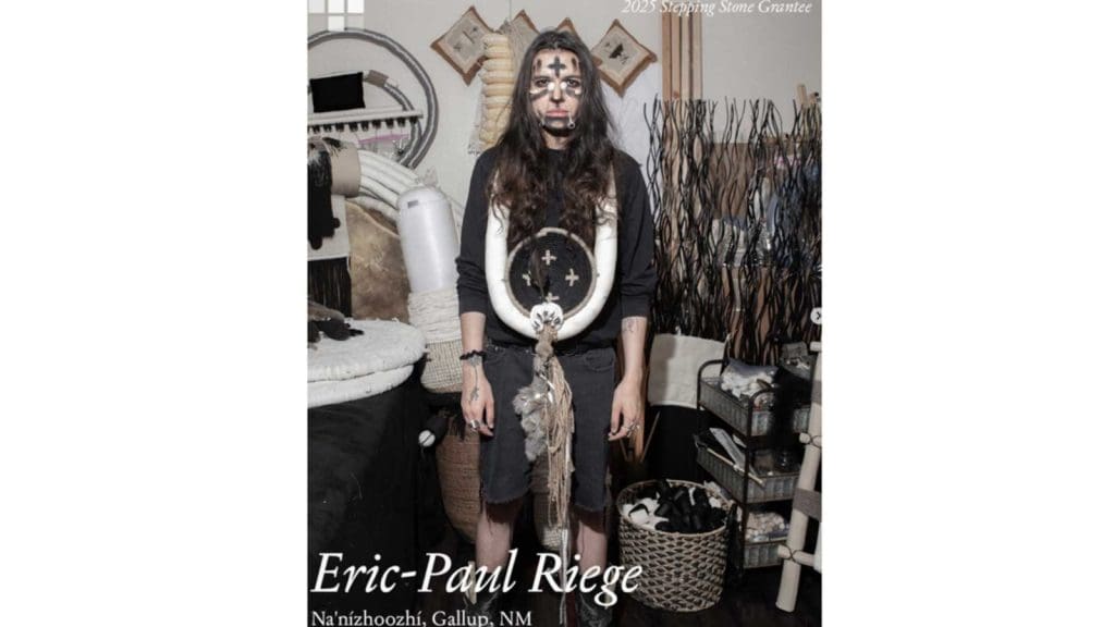 Eric-Paul Riege stands in his studio wearing woven sculptural jewelry and face paint, surrounded by fiber materials and tools.