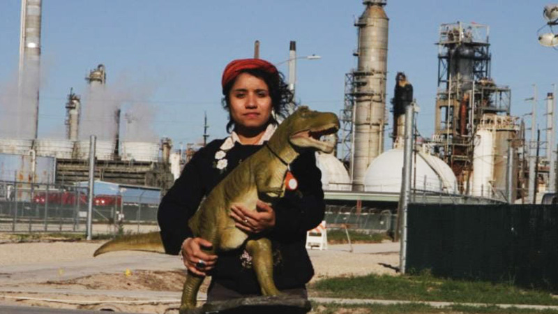 a woman holds a T-Rex doll or figure in front of oil equipment